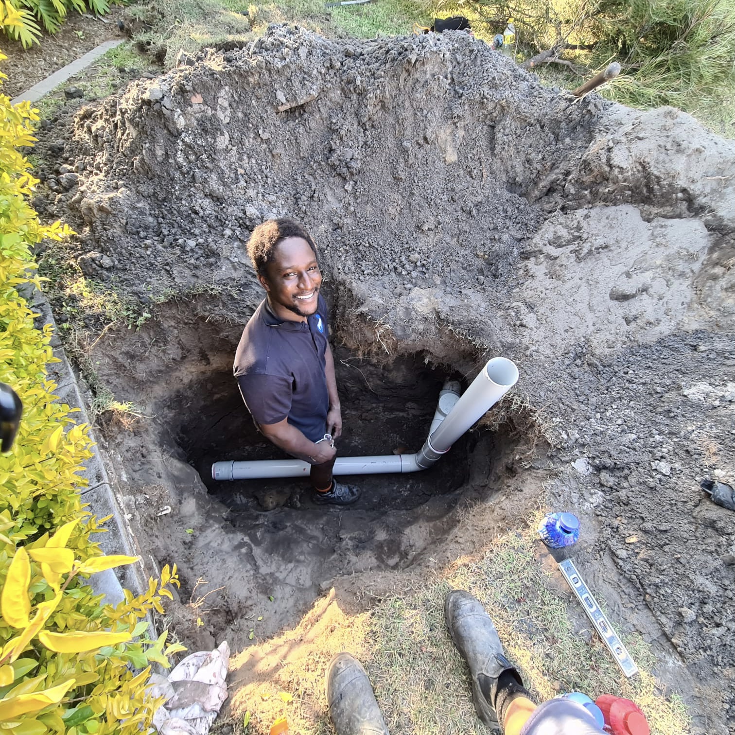 A smiling PKG Plumbing team member standing in a deep excavation beside new PVC pipework